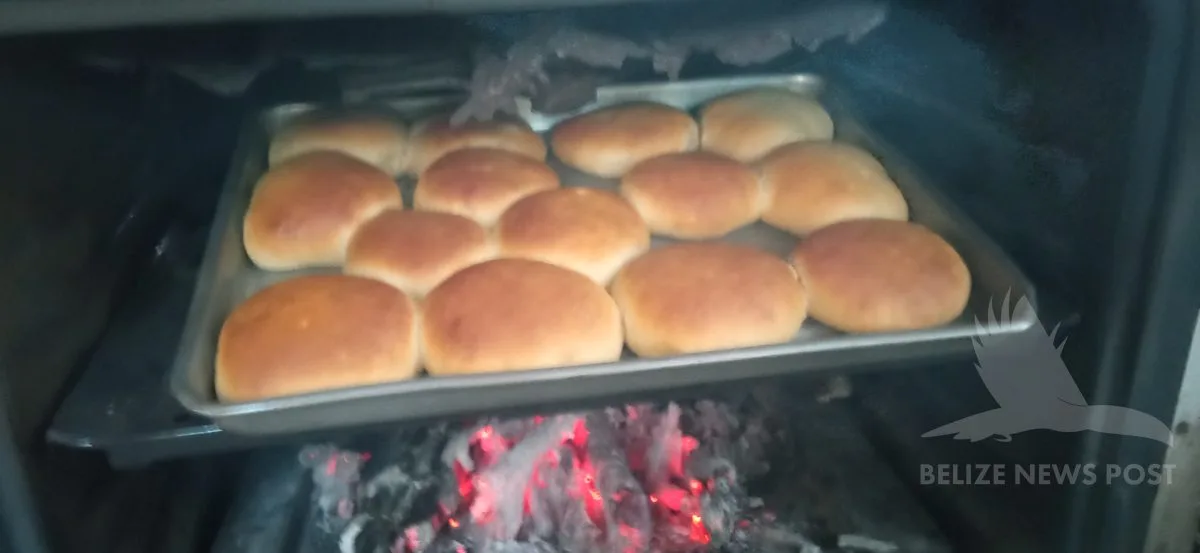 Belizean buns baking over a wood fire in a converted hearth oven, golden brown on a steel tray with glowing coals visible below