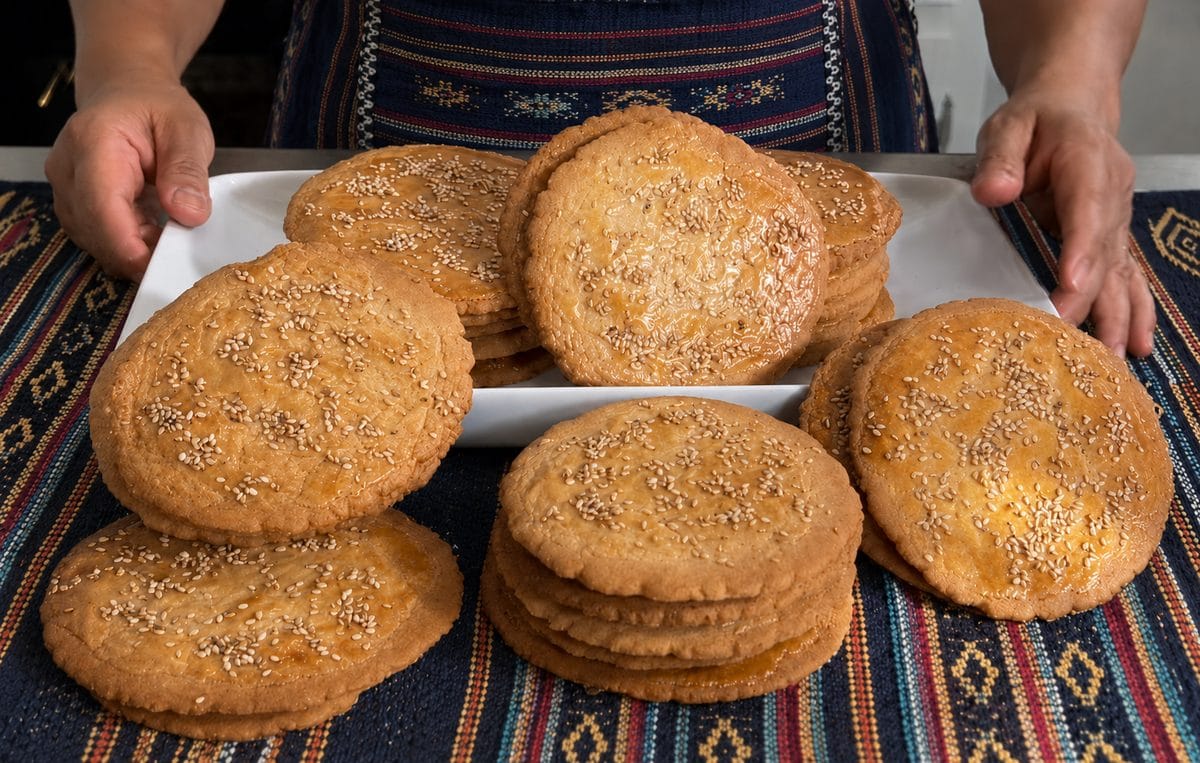 Stacks of Belizean champurradas, flat sesame-topped cookies, on a white platter over a striped woven cloth
