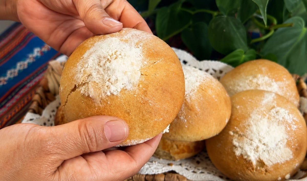 Hands holding a flour-dusted sweet bread roll over a basket of rolls, the pan de manteca base for molletes guatemaltecos