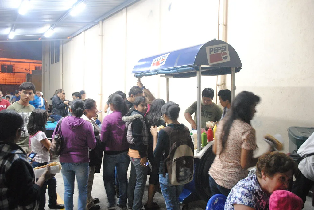 Crowd lined up at a Guatemalan shuco street cart at night with condiment bottles