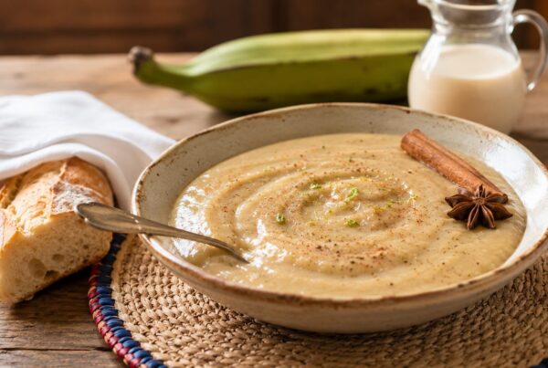 Bowl of Haitian labouyi bannann, a creamy green plantain porridge dusted with cinnamon and topped with a cinnamon stick and star anise, served with bread and evaporated milk