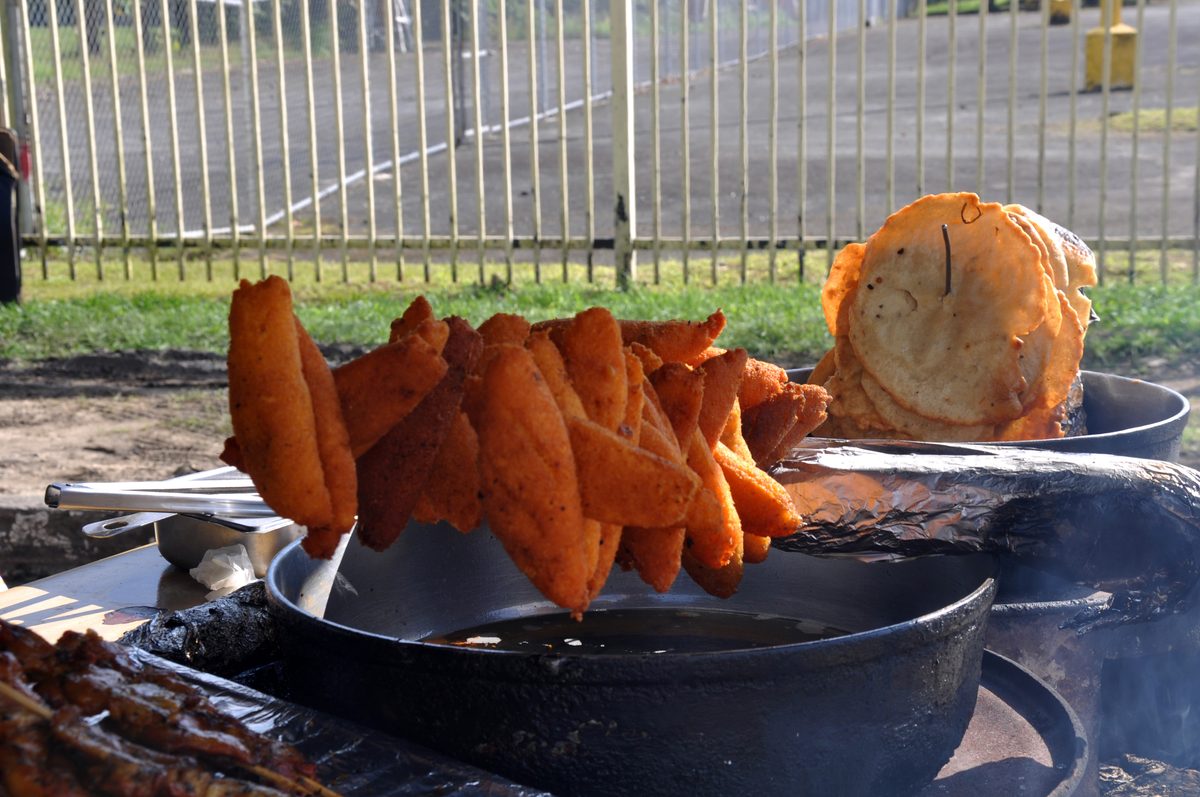 a roadside kiosk fryer with alcapurrias draining on the pot rim and a stack of flat bacalaitos behind them