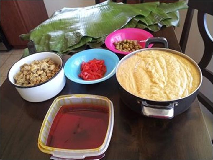 ingredients laid out for puerto rican pasteles: green banana masa in a pot, cubed pork, banana leaves, chickpeas, pimiento peppers and annatto oil