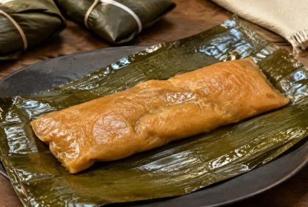 An unwrapped Puerto Rican pastel on a banana leaf, its annatto-orange masa still glossy, with two tied yuntas behind it
