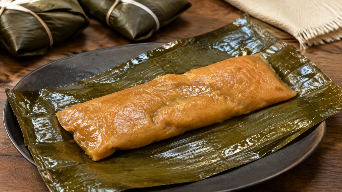 An unwrapped Puerto Rican pastel on a banana leaf, its annatto-orange masa still glossy, with two tied yuntas behind it