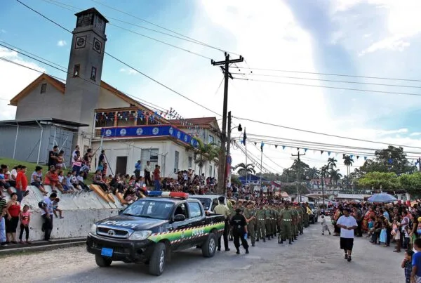 Uniformed contingent marching behind a Belize Police vehicle past buildings hung with Belize coat-of-arms bunting, with the community lining the street to watch